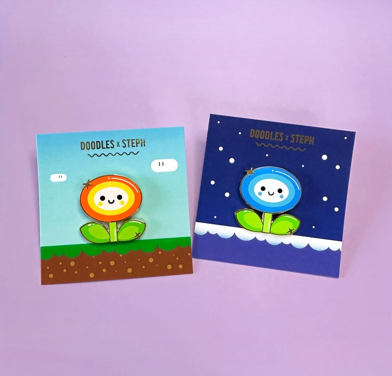 May include: Two enamel pins featuring cartoon flower designs. The first pin is a yellow and orange flower with a happy face and a star on top. The second pin is a blue flower with a happy face and a star on top. Both pins are on a white background with text that reads "DOODLES x STEPH".