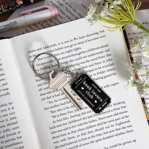 May include: A silver key ring with two keys and a black tag that reads "book lovers club" with the text "lifetime membership" and the number "0001". The keychain rests on an open book.
