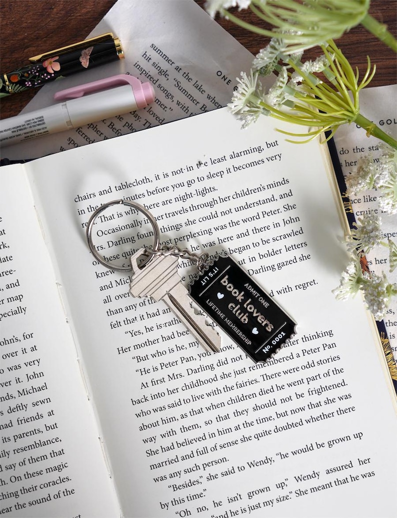 May include: A silver key ring with two keys and a black tag that reads "book lovers club" with the text "lifetime membership" and the number "0001". The keychain rests on an open book.