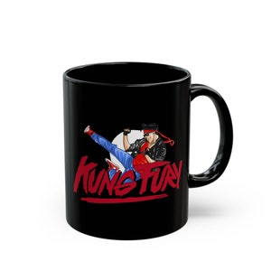 Può includere: Tazza da caffè in ceramica nera con un cerchio bianco e un personaggio dei cartoni animati che calcia in aria. Il testo "Kung Fury" è scritto in lettere rosse sotto il personaggio.