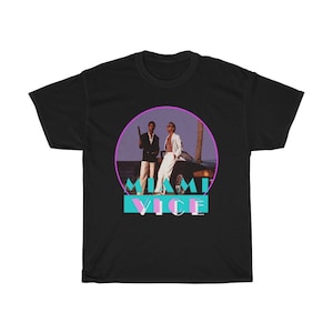 Op de afbeelding: Zwart T-shirt met een afbeelding van twee mannen die voor een auto staan. De afbeelding is in een cirkel met een roze rand. De tekst "Miami Vice" staat onder de cirkel in een blauw en roze lettertype.