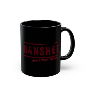 Op de afbeelding: Zwarte keramische koffiemok met een rode afbeelding waarop "Banshee" en "Small Town. Big Secrets." staat.