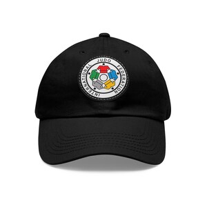 Peut inclure: Casquette de baseball noire avec un patch brodé représentant le logo de la Fédération Internationale de Judo. Le logo est un cercle avec les mots "Fédération Internationale de Judo" autour du bord et une étoile à cinq branches au centre. L'étoile est composée de cinq judogis de couleurs différentes.