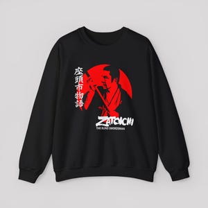 May include: A black long-sleeve sweatshirt featuring a graphic print of a person holding a sword against a large red circle. The design includes white Japanese characters and the text Zatoichi The Blind Swordsman in white lettering.