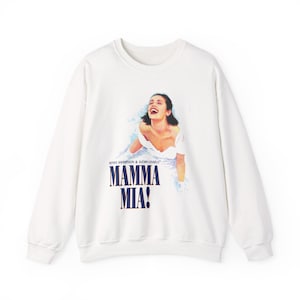 Pode incluir: Camisola branca com um gráfico do musical "Mamma Mia!". O design apresenta uma mulher em um vestido branco e o texto "MAMMA MIA!" em azul marinho. A camisola tem gola redonda e mangas compridas.