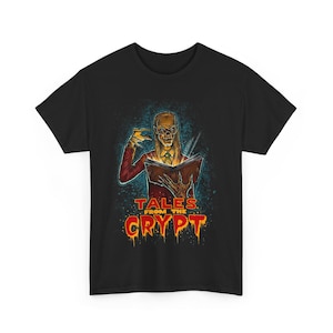 以下が含まれることがあります： 「Tales From The Crypt」のグラフィックが特徴の黒いTシャツ。赤いジャケットを着て本を持っている骸骨の姿が描かれています。「TALES FROM THE CRYPT」の文字はオレンジ色で滴り落ちるように表現されています。