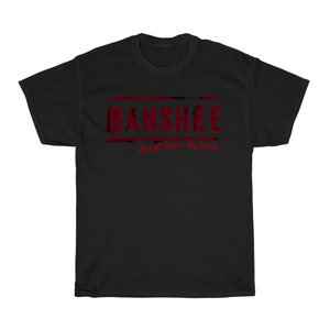 Op de afbeelding: Zwart T-shirt met een rode grafische print met de tekst "Banshee" en daaronder de tekst "Small Town. Big Secrets".