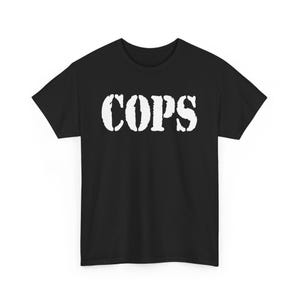 May include: Black t-shirt with the word "COPS" printed in large, distressed white letters across the chest. The shirt has short sleeves and a crew neck.