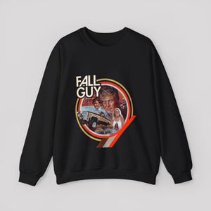 Peut inclure: Un sweat-shirt noir à manches longues avec une impression graphique circulaire portant le texte "FALL GUY". Le motif comprend le portrait d'un homme, d'une femme, d'un pick-up et d'une moto, encadrés par des lignes circulaires orange et marron.