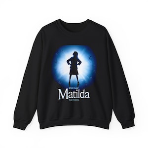 Pode incluir: Camisola preta com um gráfico azul da silhueta de uma menina e as palavras "Roald Dahl's Matilda The Musical" em branco. A camisola de manga comprida tem gola redonda.