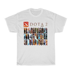 Pu&ograve; includere: T-shirt bianca con il logo "DOTA 2" in rosso e oro, e una serie di illustrazioni colorate di personaggi. Il design &egrave; stampato su una classica t-shirt girocollo.