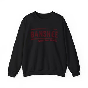 Op de afbeelding: Zwarte sweatshirt met het woord "BANSHEE" in verweerde rode letters. Daaronder staat de zin "Small Town. Big Secrets" in het rood. De sweatshirt heeft lange mouwen en een ronde hals.