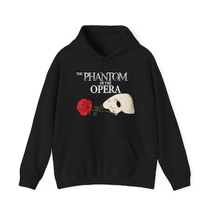 Puede incluir: Sudadera con capucha negra con el texto "The Phantom of the Opera". El diseño incluye una rosa roja y una máscara blanca. La sudadera tiene un bolsillo delantero y una capucha con cordón.