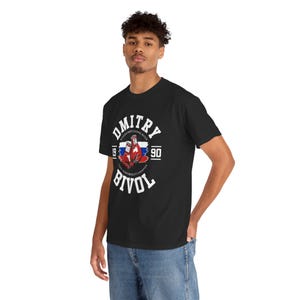 May include: Black t-shirt featuring the name "Dmitry Bivol" in white arched text. The shirt includes a graphic of red boxing gloves and the Russian flag, with the years "1990" and "90" on either side of the graphic.