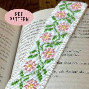 Könnte beinhalten: Ein weißes Kreuzstich-Lesezeichen mit floralem Design. Das Lesezeichen zeigt rosa Blüten mit gelben Zentren und grünen Blättern. Der Text "PDF PATTERN" befindet sich in einem rosa Oval. Das Lesezeichen befindet sich in einem offenen Buch.