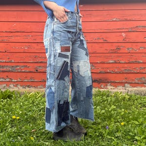 Peut inclure: Un pantalon en jean patchwork à jambes larges, dans diverses nuances de bleu. Le jean présente de multiples patchs de tissu et une étiquette en cuir marron. Le vêtement est porté à l'extérieur contre un mur en bois rouge et de l'herbe verte.