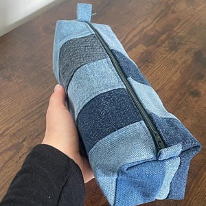 May include: A rectangular denim pencil case with a zip closure. The case is made from various shades of blue denim patches, creating a striped pattern. A small denim loop is attached to the zip end.