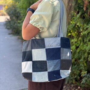 Puede incluir: Un bolso tote de mezclilla patchwork con un patrón a cuadros en tonos de azul, gris y blanco. El bolso tiene correas de mezclilla y se lleva al hombro. El fondo presenta vegetación y una pasarela pavimentada.