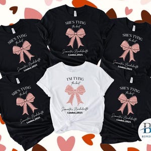 Puede incluir: Cinco camisetas negras con lazos de purpurina rosa y el texto "She's tying the knot" o "I'm tying the knot" y el nombre de la novia y la ubicación. Las camisetas son para una despedida de soltera.
