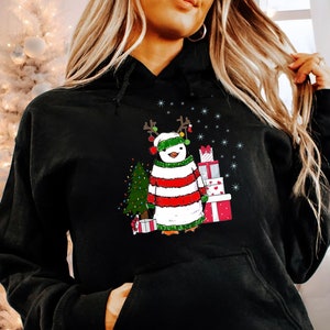 Puede incluir: Sudadera negra con un ping&uuml;ino navide&ntilde;o que lleva un su&eacute;ter a rayas rojas y blancas, un gorro verde y cuernos de reno. El ping&uuml;ino est&aacute; de pie junto a un peque&ntilde;o &aacute;rbol de Navidad y una pila de regalos envueltos.