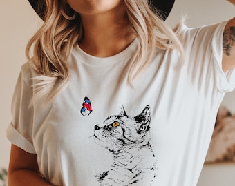 May include: White t-shirt with a black and white illustration of a cat looking up at a colorful butterfly.
