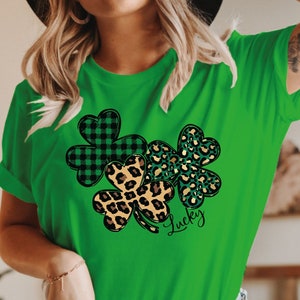 May include: Green t-shirt featuring a design of three shamrocks. The shamrocks are made from green and black checked fabric, leopard print, and green and gold glitter. The word "Lucky" is written beneath the shamrocks.