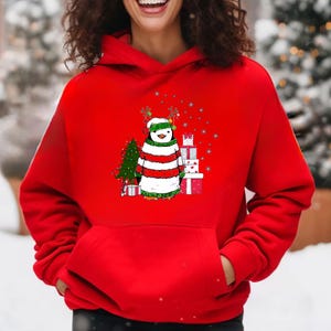 Puede incluir: Sudadera roja con capucha con un ping&uuml;ino navide&ntilde;o que lleva un su&eacute;ter a rayas y cuernos de reno. El ping&uuml;ino est&aacute; de pie junto a un peque&ntilde;o &aacute;rbol de Navidad y regalos.