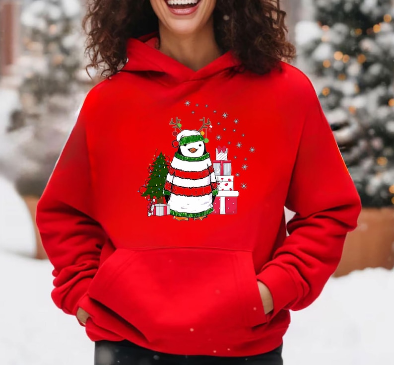 Puede incluir: Sudadera roja con capucha con un ping&uuml;ino navide&ntilde;o que lleva un su&eacute;ter a rayas y cuernos de reno. El ping&uuml;ino est&aacute; de pie junto a un peque&ntilde;o &aacute;rbol de Navidad y regalos.