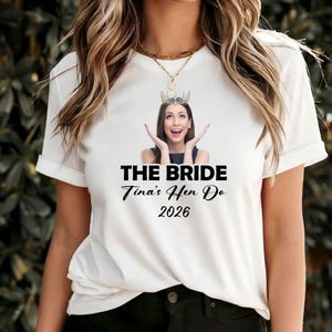 May include: A white short-sleeved t-shirt featuring a graphic of a person wearing a crown with hands raised near their face. Below the image, the text reads THE BRIDE, Tina's Hen Do, and 2026 in black lettering. The shirt has a crew neckline.