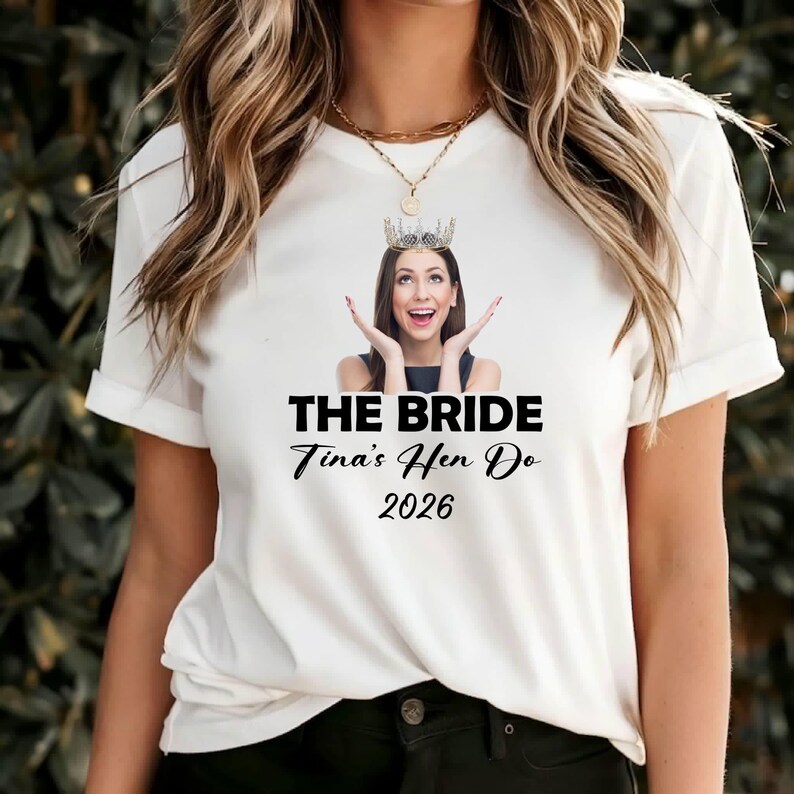 May include: A white short-sleeved t-shirt featuring a graphic of a person wearing a crown with hands raised near their face. Below the image, the text reads THE BRIDE, Tina's Hen Do, and 2026 in black lettering. The shirt has a crew neckline.