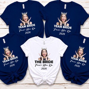 May include: Four navy blue t-shirts and one white t-shirt, each featuring a custom photo of a person wearing a crown, with the text TEAM BRIDE or THE BRIDE, Tina's Hen Do, and 2026 printed in white or black lettering on the front of the shirts.