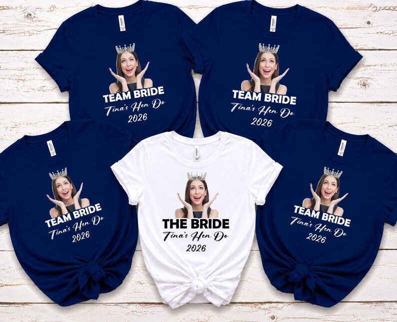 May include: Four navy blue t-shirts and one white t-shirt, each featuring a custom photo of a person wearing a crown, with the text TEAM BRIDE or THE BRIDE, Tina's Hen Do, and 2026 printed in white or black lettering on the front of the shirts.