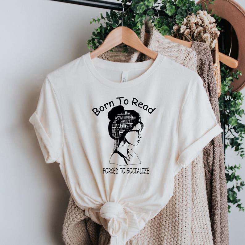 May include: A white t-shirt with a black graphic of a woman with a stack of books for a head. The text "Born to Read Forced to Socialize" is printed on the shirt.