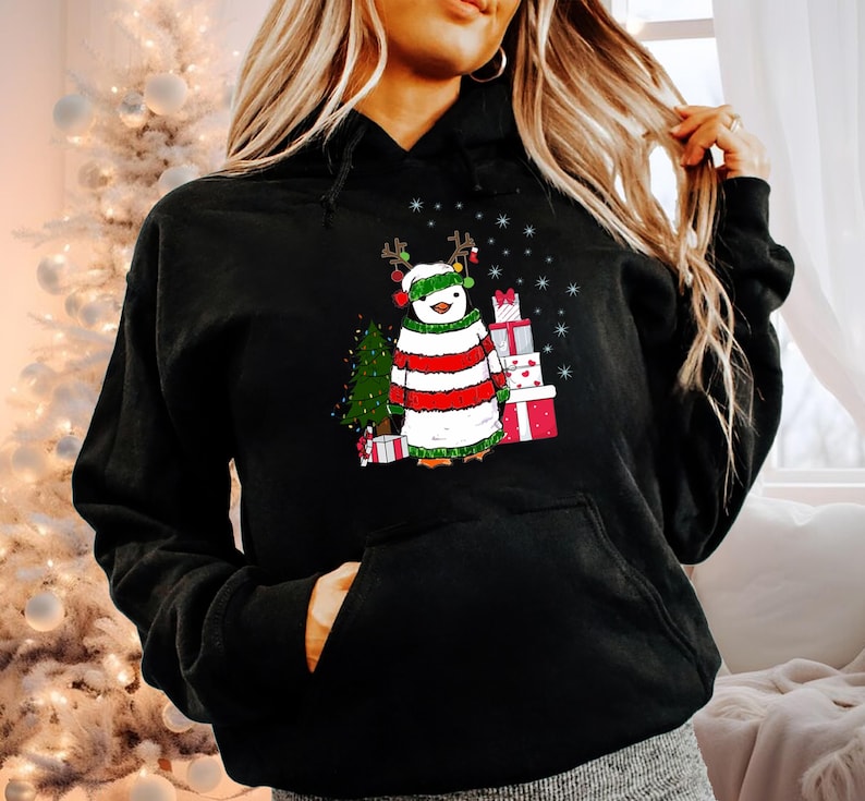Puede incluir: Sudadera negra con un ping&uuml;ino navide&ntilde;o que lleva un su&eacute;ter a rayas rojas y blancas, un gorro verde y cuernos de reno. El ping&uuml;ino est&aacute; de pie junto a un peque&ntilde;o &aacute;rbol de Navidad y una pila de regalos envueltos.