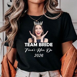 May include: A black short-sleeved t-shirt featuring a graphic of a person wearing a crown, with the text "TEAM BRIDE", "Tina's Hen Do", and "2026" printed in white below the image. The shirt has a crew neckline and a standard fit design.