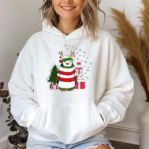 Puede incluir: Sudadera con capucha blanca con un ping&uuml;ino navide&ntilde;o que lleva un su&eacute;ter a rayas rojas y verdes y un gorro de Pap&aacute; Noel. El ping&uuml;ino sostiene un regalo y est&aacute; de pie frente a un peque&ntilde;o &aacute;rbol de Navidad.