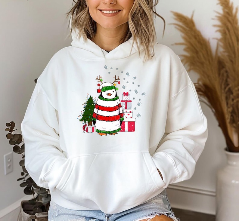 Puede incluir: Sudadera con capucha blanca con un ping&uuml;ino navide&ntilde;o que lleva un su&eacute;ter a rayas rojas y verdes y un gorro de Pap&aacute; Noel. El ping&uuml;ino sostiene un regalo y est&aacute; de pie frente a un peque&ntilde;o &aacute;rbol de Navidad.