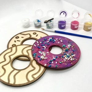 Peut inclure: Kit de peinture de donuts avec trois formes de donuts en bois, des pots de peinture, des paillettes et un pinceau. Un donut est peint en violet avec des paillettes, les autres sont non peints avec des motifs gravés.