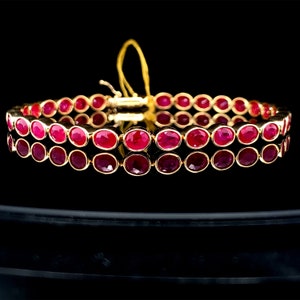 May include: A gold bracelet with two rows of oval-cut rubies. The rubies are a deep red hue and are set in a gold bezel. The bracelet has a clasp and is shown on a reflective black surface. A luxury jewellery item.