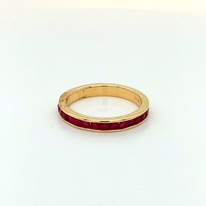May include: A gold band ring with a row of square-cut, vibrant red gemstones. The ring is set against a white background, highlighting the contrast between the gold and the red stones. The ring's design is simple and elegant.