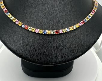 May include: A gold-toned necklace with a row of colorful gemstones. The gemstones are in various colors, including red, pink, yellow, blue, and green. The necklace is displayed on a black jewelry stand.