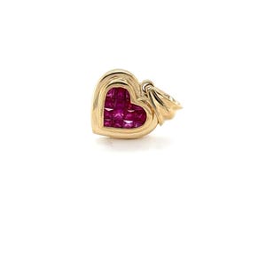 May include: A gold heart-shaped pendant featuring a cluster of square-cut, deep red gemstones. The pendant has a rounded, polished gold frame and a small loop for a chain. The gemstones fill the heart shape, creating a vibrant contrast against the gold.