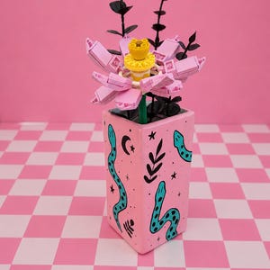 May include: A square pink ceramic vase decorated with black hand-painted snakes, stars, and foliage motifs. The vase contains a constructed plastic building block flower with light pink petals and a yellow centre, accompanied by black artificial stems.