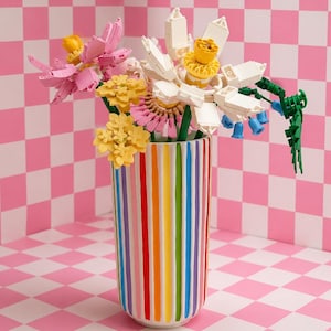 May include: A cylindrical ceramic vase featuring vertical stripes in rainbow colours including red, orange, yellow, green, blue, and purple. The vase holds a bouquet of assorted artificial flowers constructed from plastic building blocks in pink, white, and yellow.