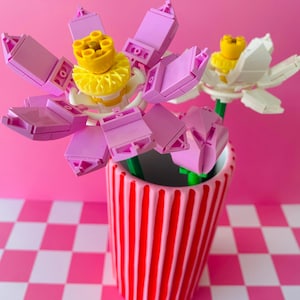 May include: A decorative vase with Lego flowers. The vase is red with white stripes. The flowers are made from pink, yellow, and white Lego bricks, with green stems. The background is pink, and the surface is a pink and white checkered pattern.