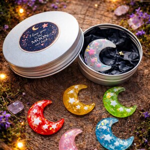 May include: A selection of colourful crescent moon-shaped resin charms with embedded stars. One is in a silver tin with the text "I love you to the MOON and back." The moons are red, yellow, pink, green, and blue.