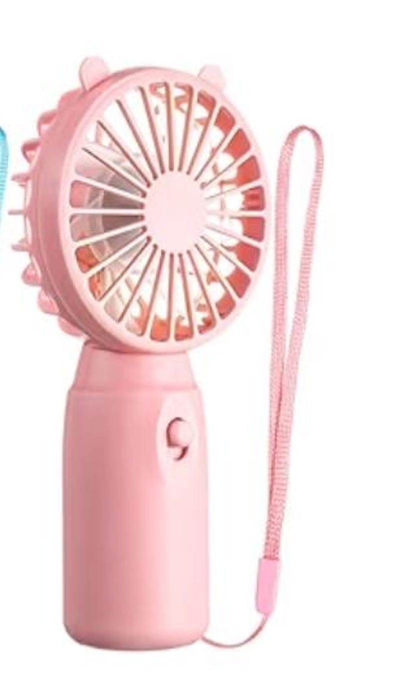 Op de afbeelding: Een roze draagbare ventilator met een bijpassende polsband. De ventilator heeft een ronde kop met een beschermend rooster en een centrale knop. Het ontwerp van de ventilator omvat kleine, oorachtige uitsteeksels aan de bovenkant.