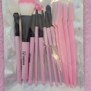 Op de afbeelding: Een doorzichtige plastic verpakking met een set roze en zwarte make-up kwasten en roze wenkbrauw scheermesjes. De kwasten hebben roze handvatten met de merknaam "VANDER". De borstelkoppen zijn wit en roze.