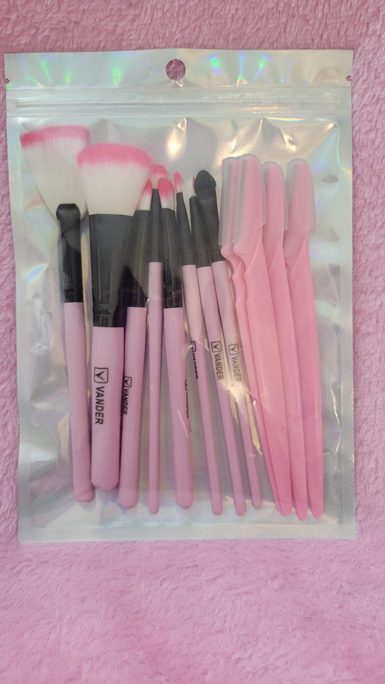 Op de afbeelding: Een doorzichtige plastic verpakking met een set roze en zwarte make-up kwasten en roze wenkbrauw scheermesjes. De kwasten hebben roze handvatten met de merknaam "VANDER". De borstelkoppen zijn wit en roze.