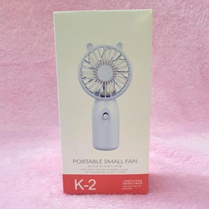 Op de afbeelding: Een witte draagbare kleine ventilator met een beren-vormige bovenkant, verpakt in een witte doos. De ventilator heeft een ronde kop met bladen en een handvat. De doos heeft de tekst "PORTABLE SMALL FAN" en "K-2".